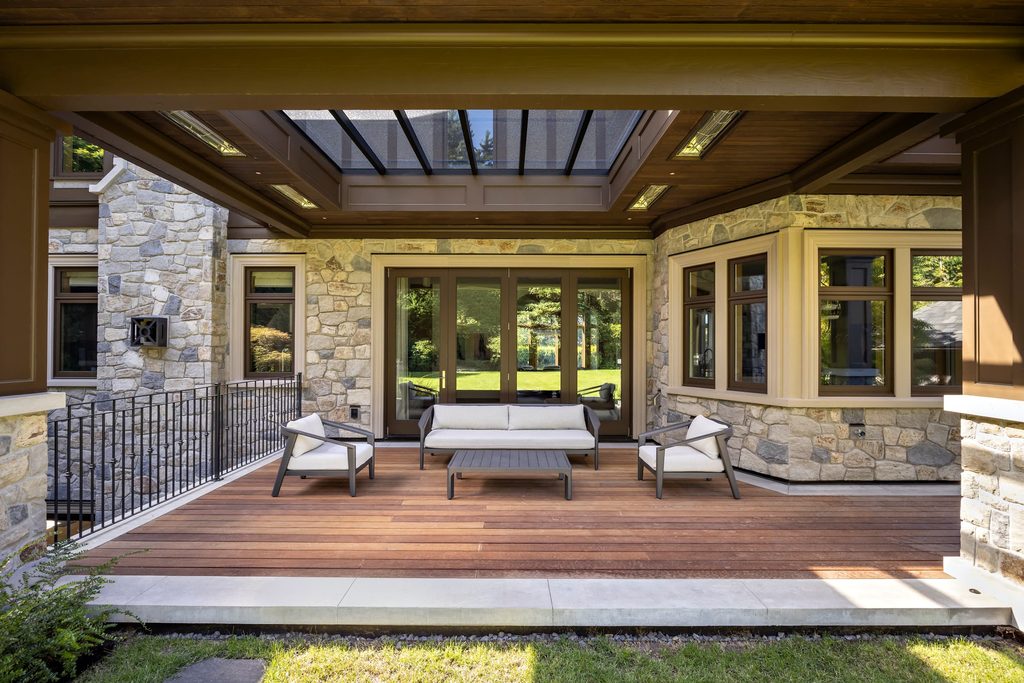 Stone-clad house exterior with a covered wooden deck and outdoor seating facing glass doors
