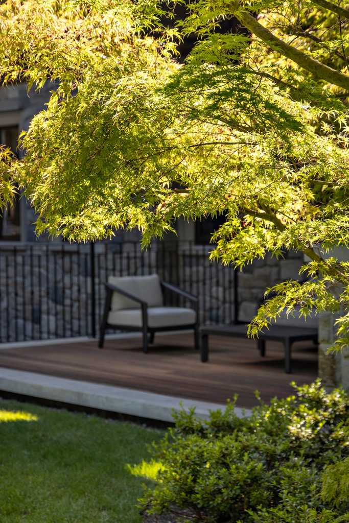 Patio seating area shaded by a lush green maple tree with two dark frame chairs on a wooden deck behind a black railing.