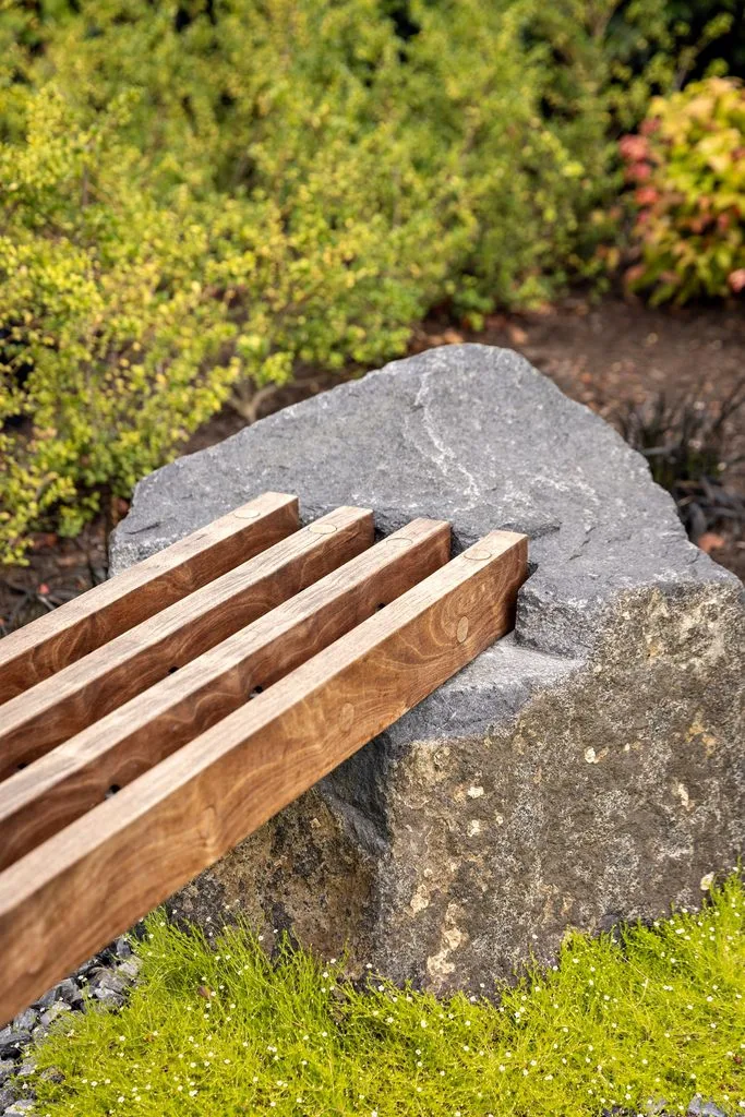 Swicks_4280_River_Web-13-8002 - Swick's Landscaping Ltd. Wooden slatted bench placed over a large gray rock in a garden with green shrubs in the background