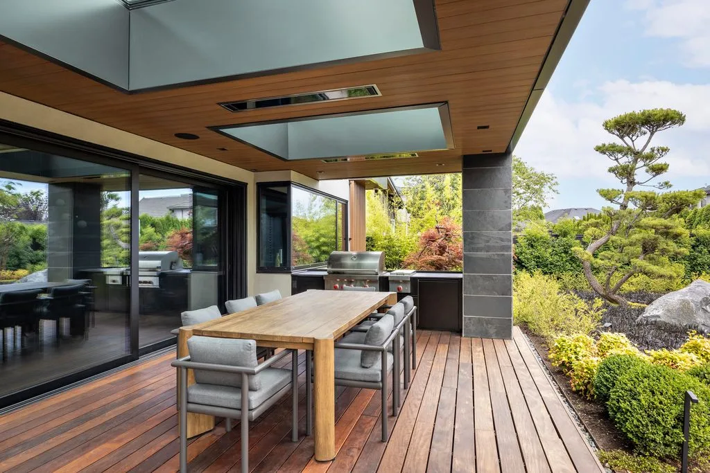 Swicks_4280_River_Web-17-2447 - Swick's Landscaping Ltd. Outdoor dining area on a wooden deck with a long table and grey cushioned chairs beside glass doors and a built-in grill.
