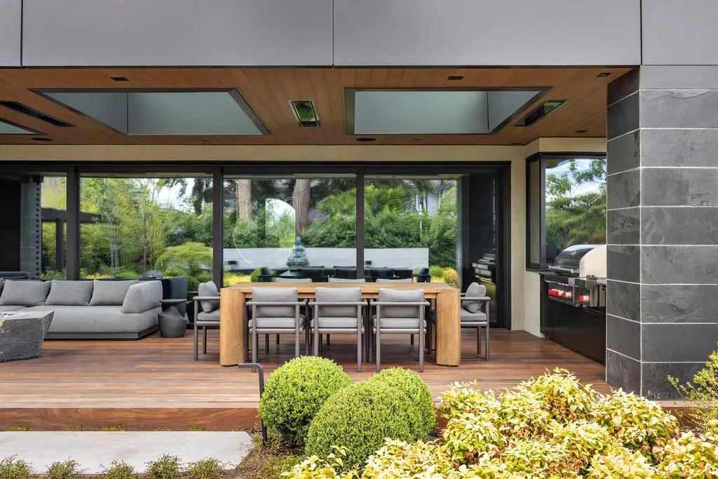 Swicks_4280_River_Web-19-2461 - Swick's Landscaping Ltd. Modern backyard patio with a wooden deck, gray sectional seating, a long wooden dining table, and a built-in grill on the right.