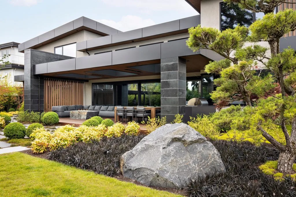 Swicks_4280_River_Web-20-2464 - Swick's Landscaping Ltd. Modern contemporary house exterior with a covered patio, gray metal accents, and large windows, surrounded by landscaped shrubs and trees outside. Engaging outdoor seating area visible under the deck.
