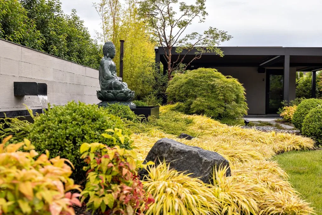 Swicks_4280_River_Web-22-8013 - Swick's Landscaping Ltd. Bronze Buddha statue in a tranquil garden with yellow and green plants and a modern building in the background down the wall fountain area.
