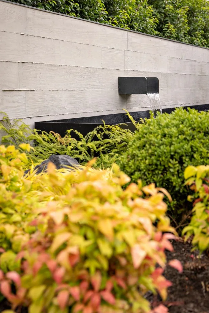 Swicks_4280_River_Web-23-8016 - Swick's Landscaping Ltd. Water feature: dark rectangular spout pouring water over a pale concrete wall with lush greenery and autumn-colored shrubs in the foreground.
