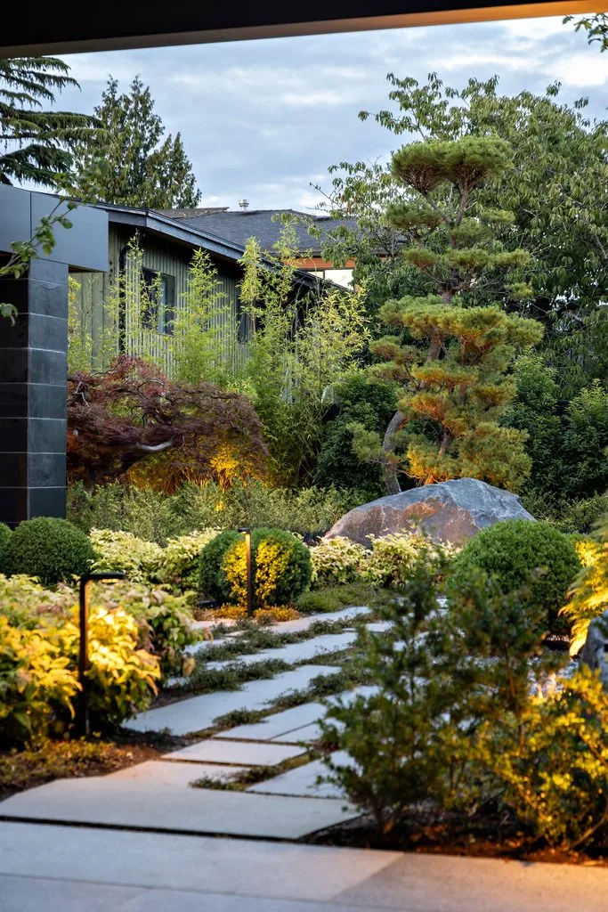 Swicks_4280_River_Web-46-8125 - Swick's Landscaping Ltd. Modern garden with stepping-stone path, low landscape lights, and neatly trimmed shrubs leading to a dark house amid trees (evening scene).