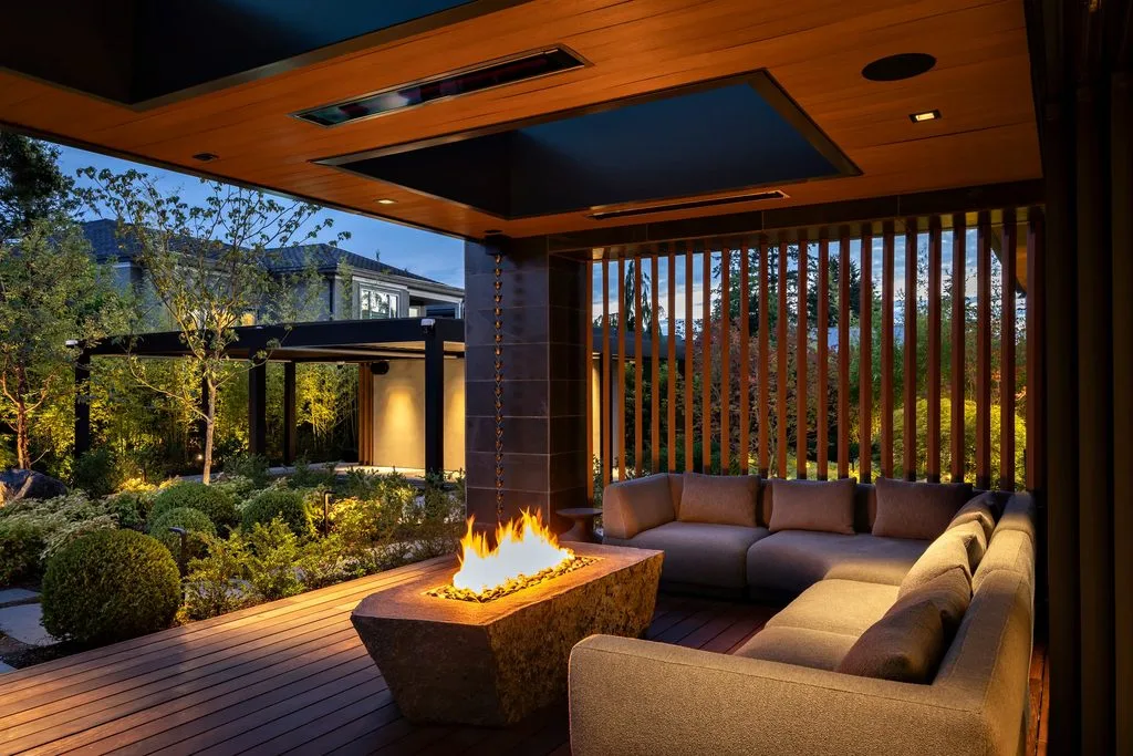 Swicks_4280_River_Web-49-2555 - Swick's Landscaping Ltd. Modern outdoor seating area at dusk with a rectangular stone fire pit, beige sectional sofa, wooden deck and vertical slat privacy screen surrounding a landscaped garden.