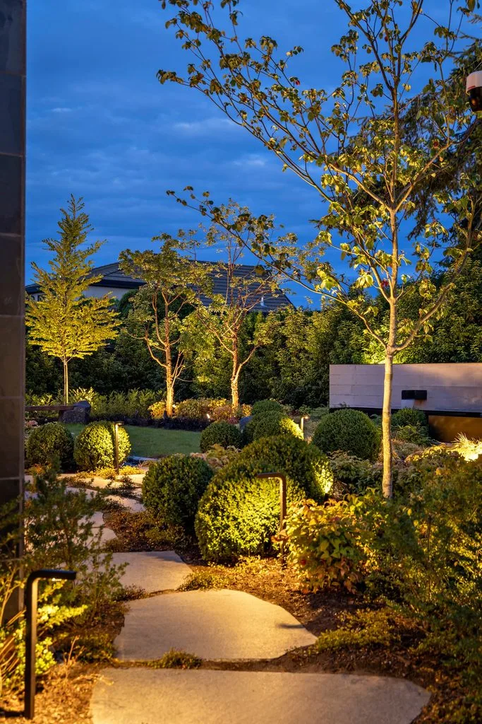 Swicks_4280_River_Web-57-8166 - Swick's Landscaping Ltd. Evening garden path lined with trimmed bushes and tall trees, illuminated by warm ground lights under a blue sky