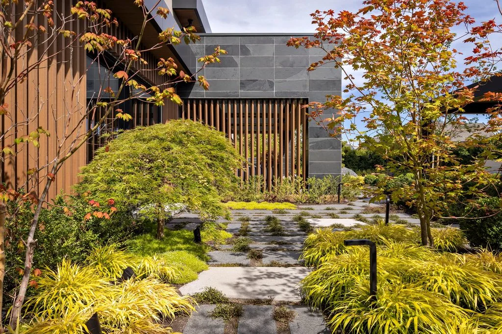 Swicks_4280_River_Web-6-978 - Swick's Landscaping Ltd. Modern building facade with gray stone wall and vertical wooden slats, surrounded by autumn‑coloured trees and yellow ornamental grasses along a stone pathway.
