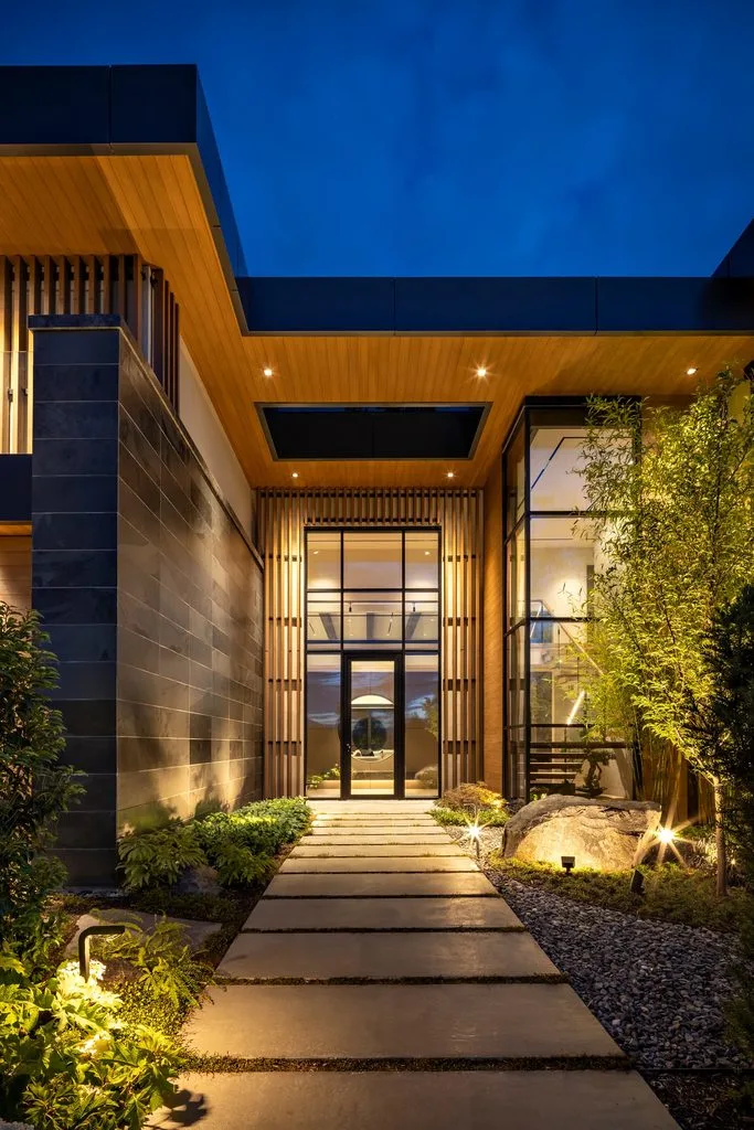 Swicks_4280_River_Web-63-8179 - Swick's Landscaping Ltd. Modern building entrance at dusk with wood ceiling, glass doors, and stone walls; landscaped path with stepping stones and gravel leading to the doorway
