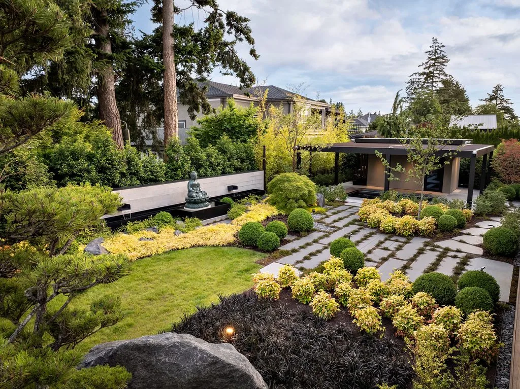 Swicks_4280_River_Web-69-0067 - Swick's Landscaping Ltd. Zen-style backyard garden with a stone Buddha statue fountain, trimmed shrubs, and paved stepping-stone paths near a modern house.