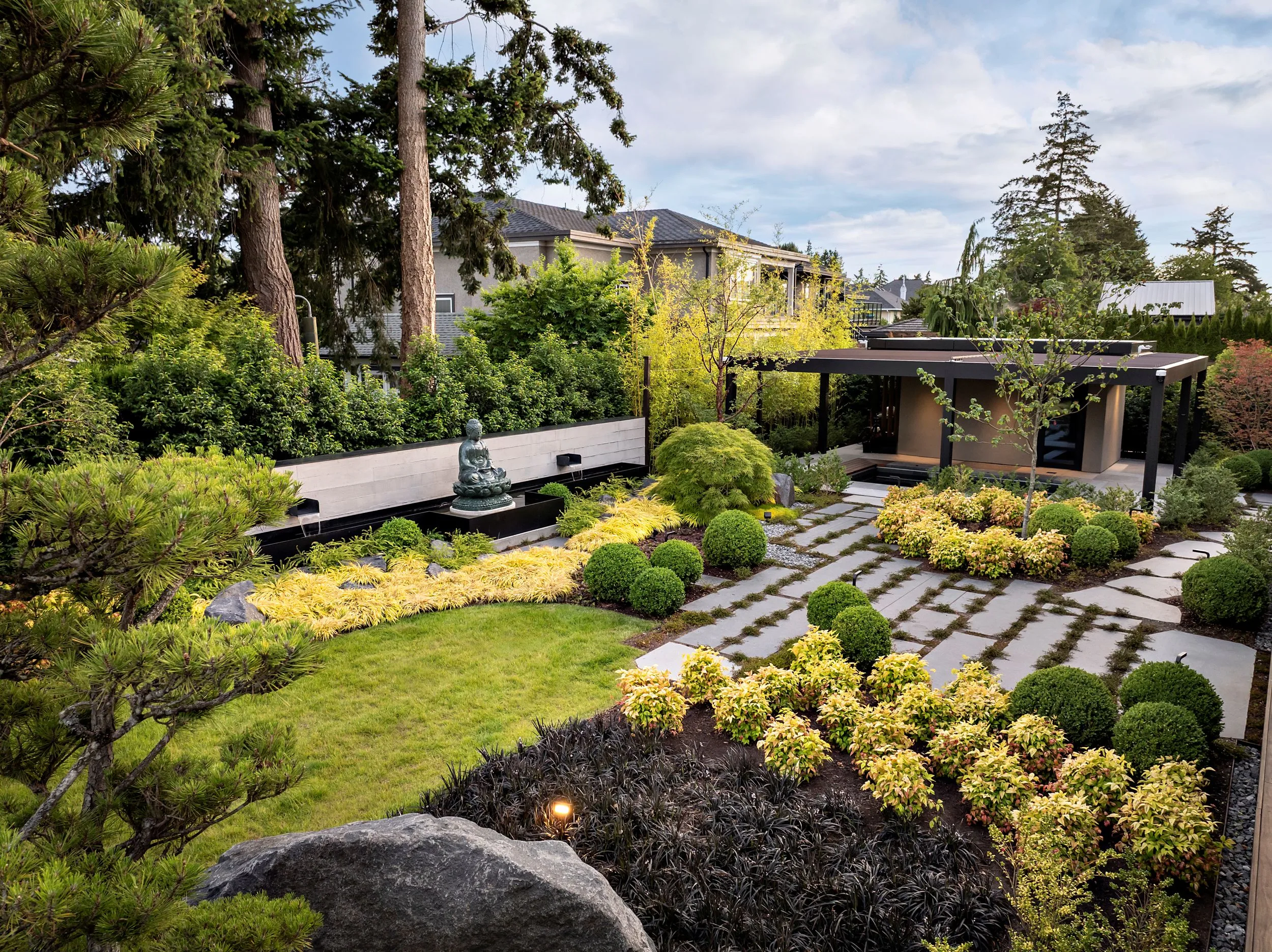 Well-kept backyard garden with ornamental shrubs, a Buddha statue fountain, and a modern house in the background.