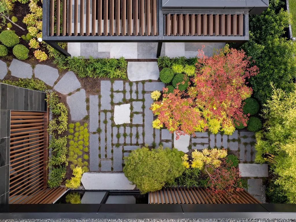 Swicks_4280_River_Web-70-0104 - Swick's Landscaping Ltd. Bird's-eye view of a modern courtyard with rectangular stepping stones, low hedges, and colorful shrubs framed by dark metal building with vertical wooden slats.