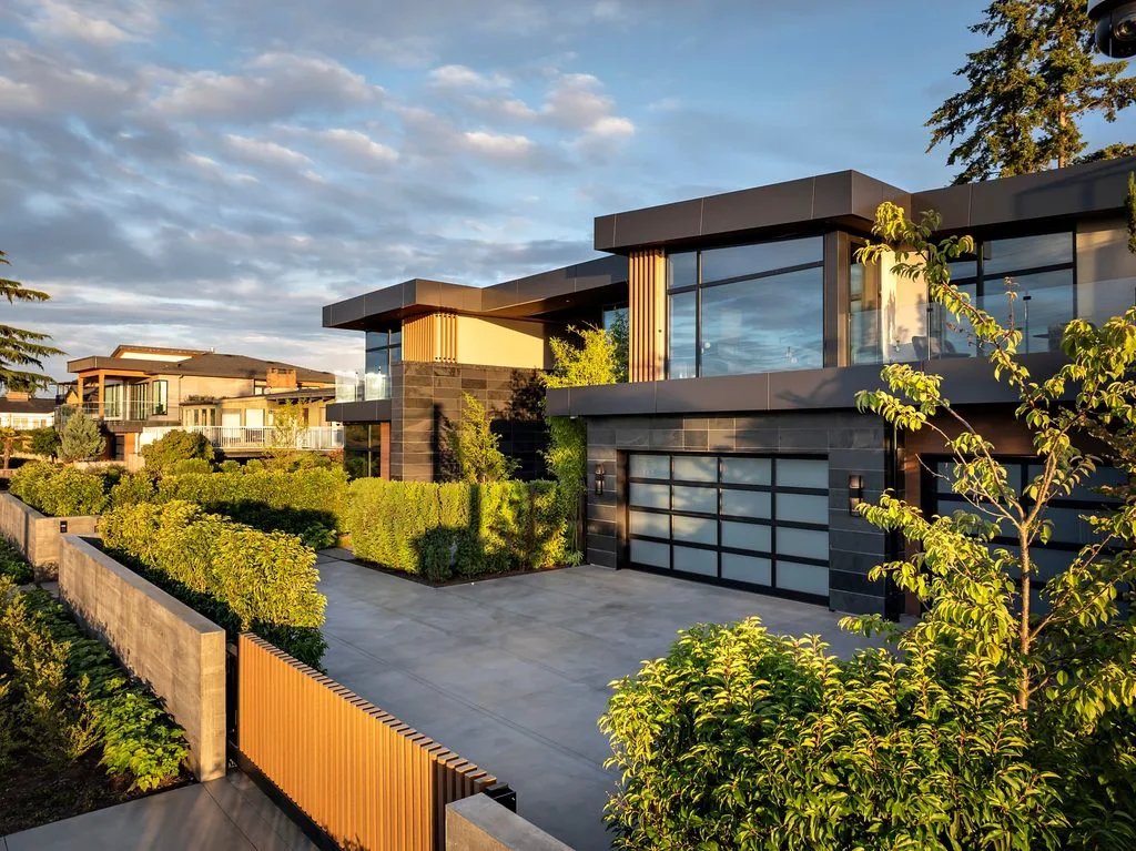 Swicks_4280_River_Web-77-0286 - Swick's Landscaping Ltd. Modern two-story house with dark grey panels, large glass windows, and orange wooden slats, surrounded by hedges and trees at golden hour.