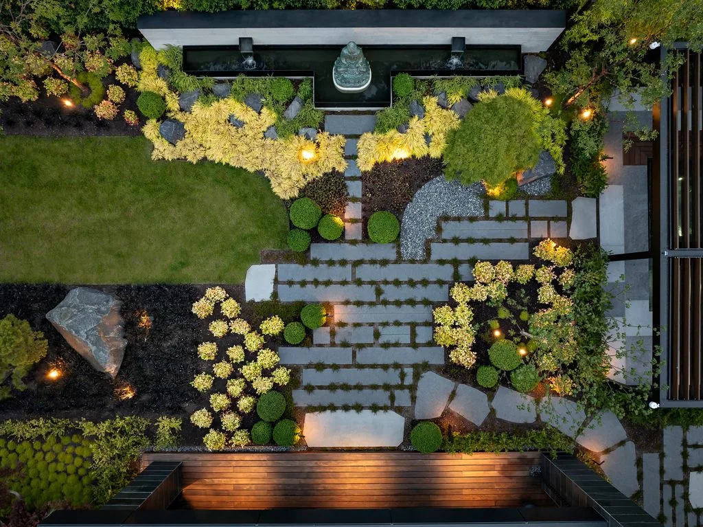 Swicks_4280_River_Web-79-0316 - Swick's Landscaping Ltd. Aerial view of a modern courtyard garden with a central water feature and rectangular stepping-stone paths surrounded by green shrubs and yellow plants at dusk