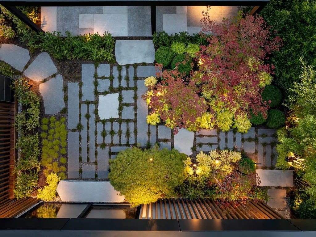 Swicks_4280_River_Web-83-0346 - Swick's Landscaping Ltd. Bird’s-eye view of a modern courtyard garden with rectangular stone stepping stones, low hedges, and colorful trees lit by warm outdoor lights.
