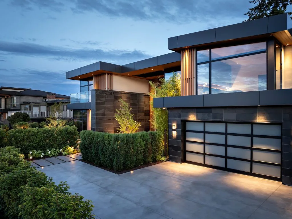 Swicks_4280_River_Web-87-0418 - Swick's Landscaping Ltd. Modern two-story house with large glass windows, dark gray panels, and a landscaped front yard at dusk, glowing outdoor lights nearby.