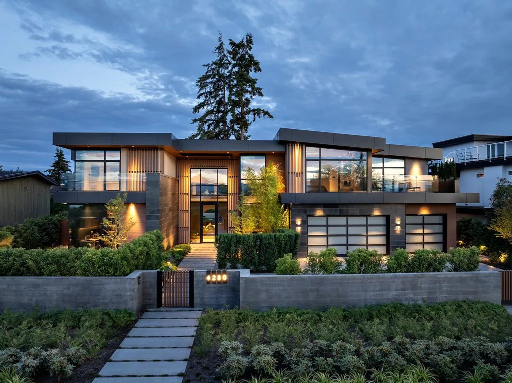 Swicks_4280_River_Web-88-0428 - Swick's Landscaping Ltd. Front view of a modern two-story house with large glass walls, warm interior lighting, and a manicured front garden under a cloudy evening sky. The gate shows the number 4280.