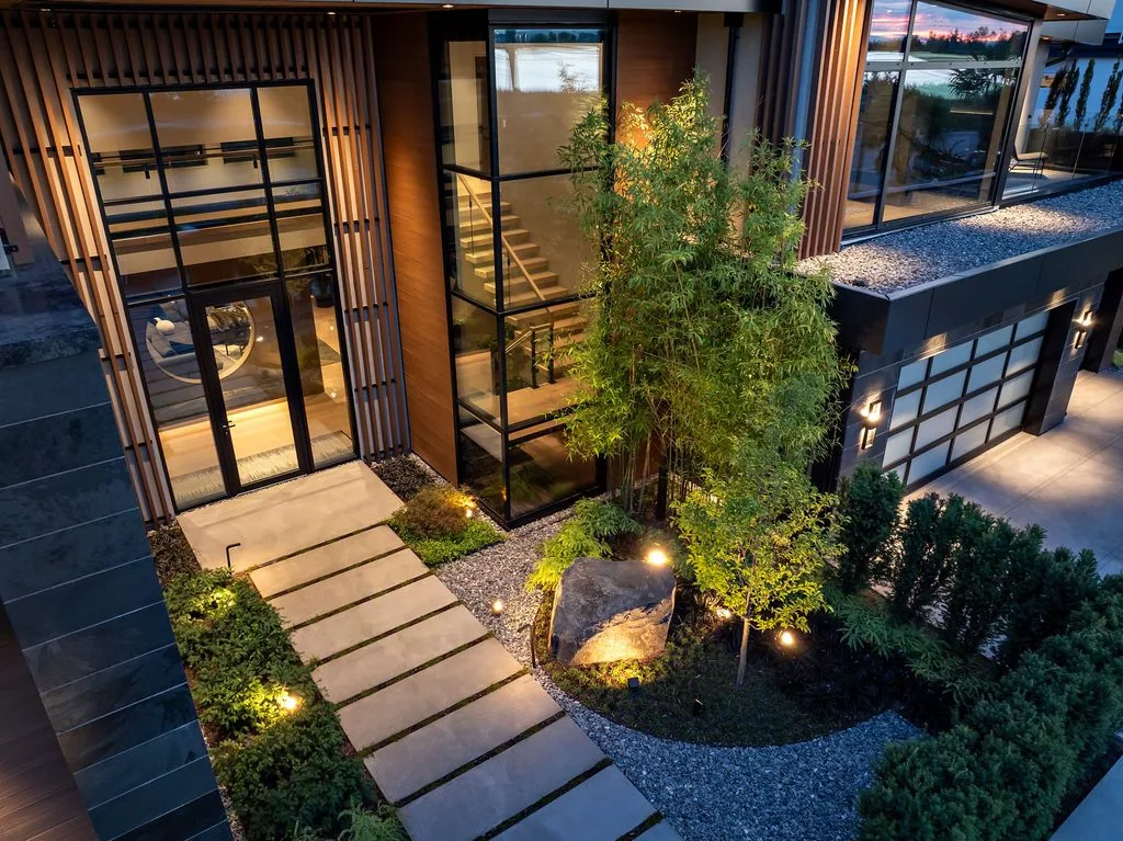 Swicks_4280_River_Web-89-0438 - Swick's Landscaping Ltd. Modern building exterior with large glass walls, vertical wooden slats, and a landscaped courtyard featuring a stepping-stone path and gravel beds at dusk.