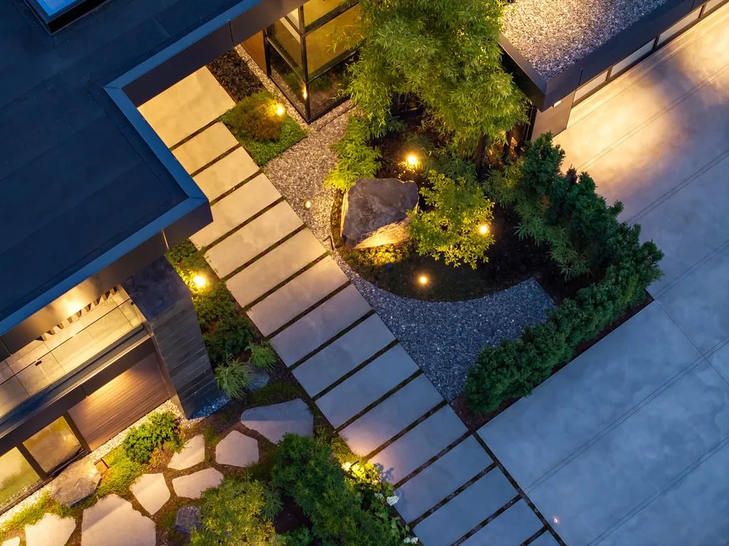 Swicks_4280_River_Web-90-0446 - Swick's Landscaping Ltd. Aerial view of a modern courtyard with rectangular stepping-stone path, gravel, and green trees lit by warm ground lights at dusk.