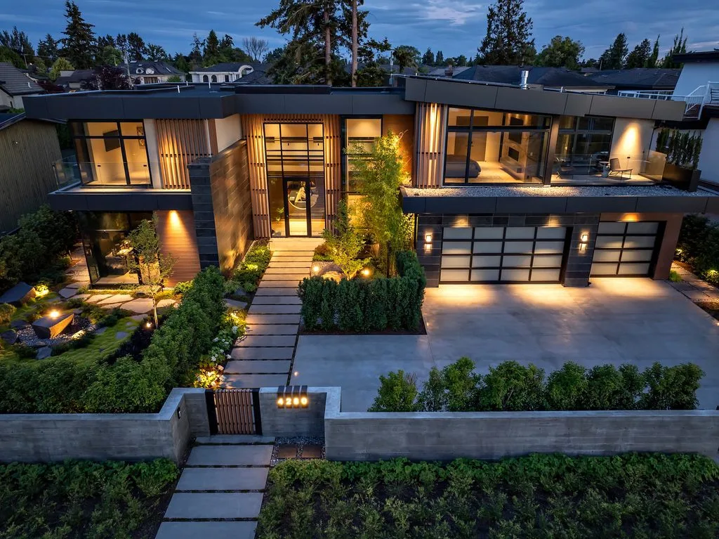Swicks_4280_River_Web-92-0483 - Swick's Landscaping Ltd. Aerial view of a modern two‑story house with glass walls, wooden slats, and warm interior lighting at dusk, surrounded by a landscaped yard and stepping stone path.